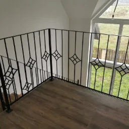 Czarna, metalowa balustrada wewnętrzna z dekoracyjnymi rombami na drewnopodobnej podłodze, widok z góry na okno z widokiem na ogród.