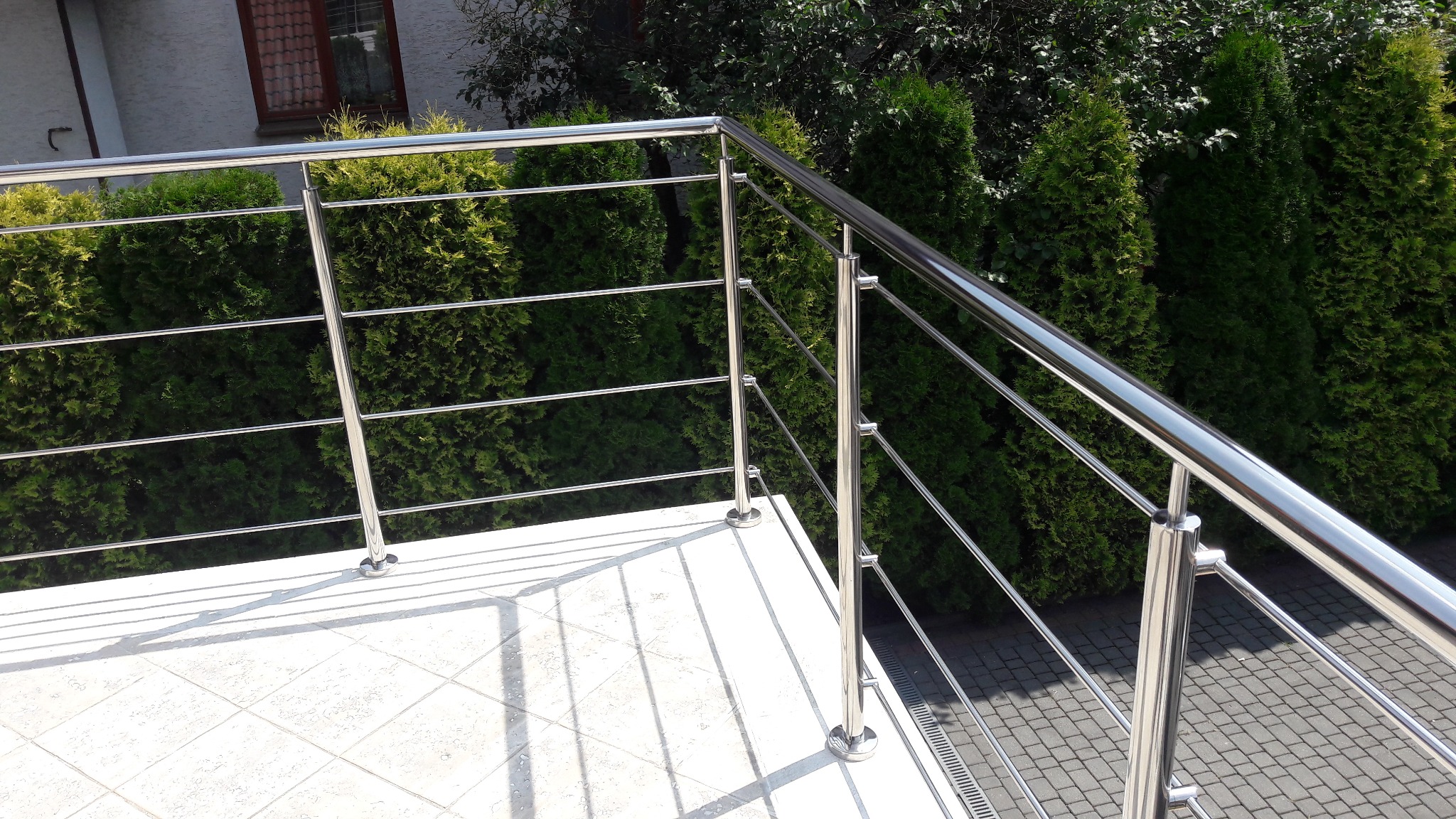 Błyszcząca, metalowa barierka balkonowa z cienkimi, poziomymi prętami, na tle zielonych krzewów i jasnej, kwadratowej płytki podłogowej, z widocznymi cieniami prętów.