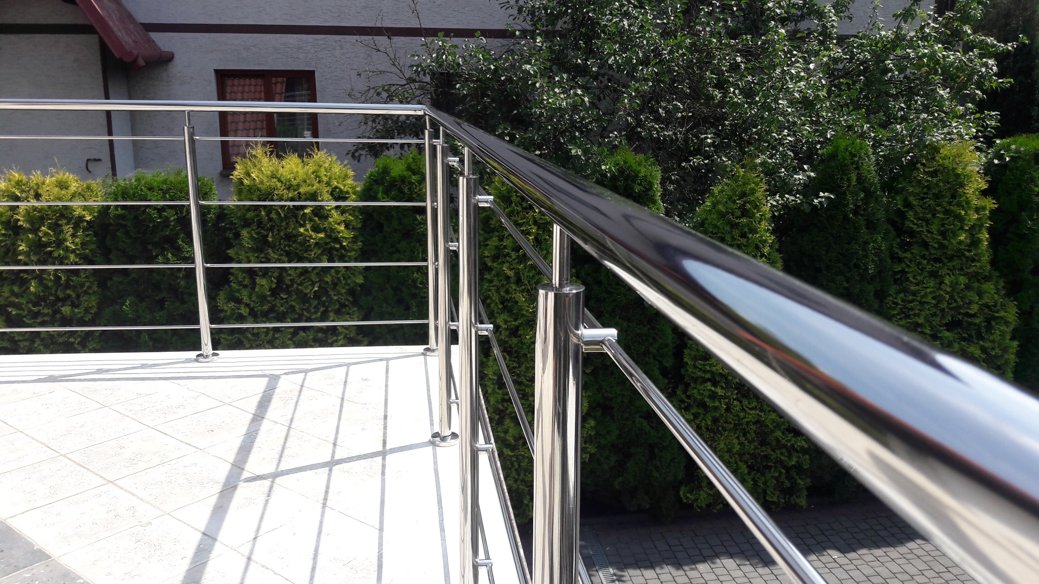 Błyszcząca, metalowa konstrukcja zabezpieczająca balkon, z widocznymi elementami mocującymi i tłem z zielonych krzewów.