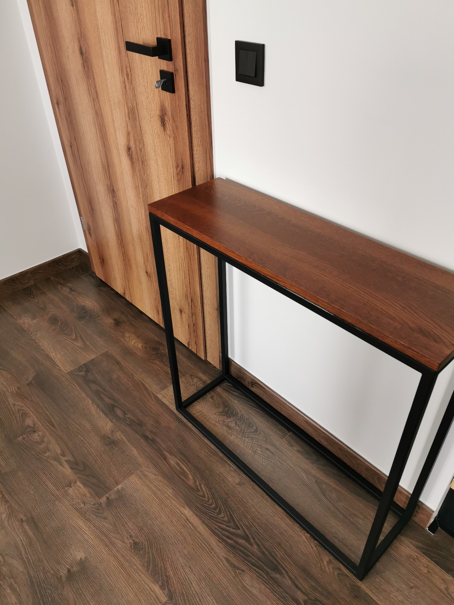 Wąski, drewniany stolik konsolowy z czarną, metalową ramą, ustawiony przy ścianie obok drewnianych drzwi z czarną klamką i włącznikiem światła.