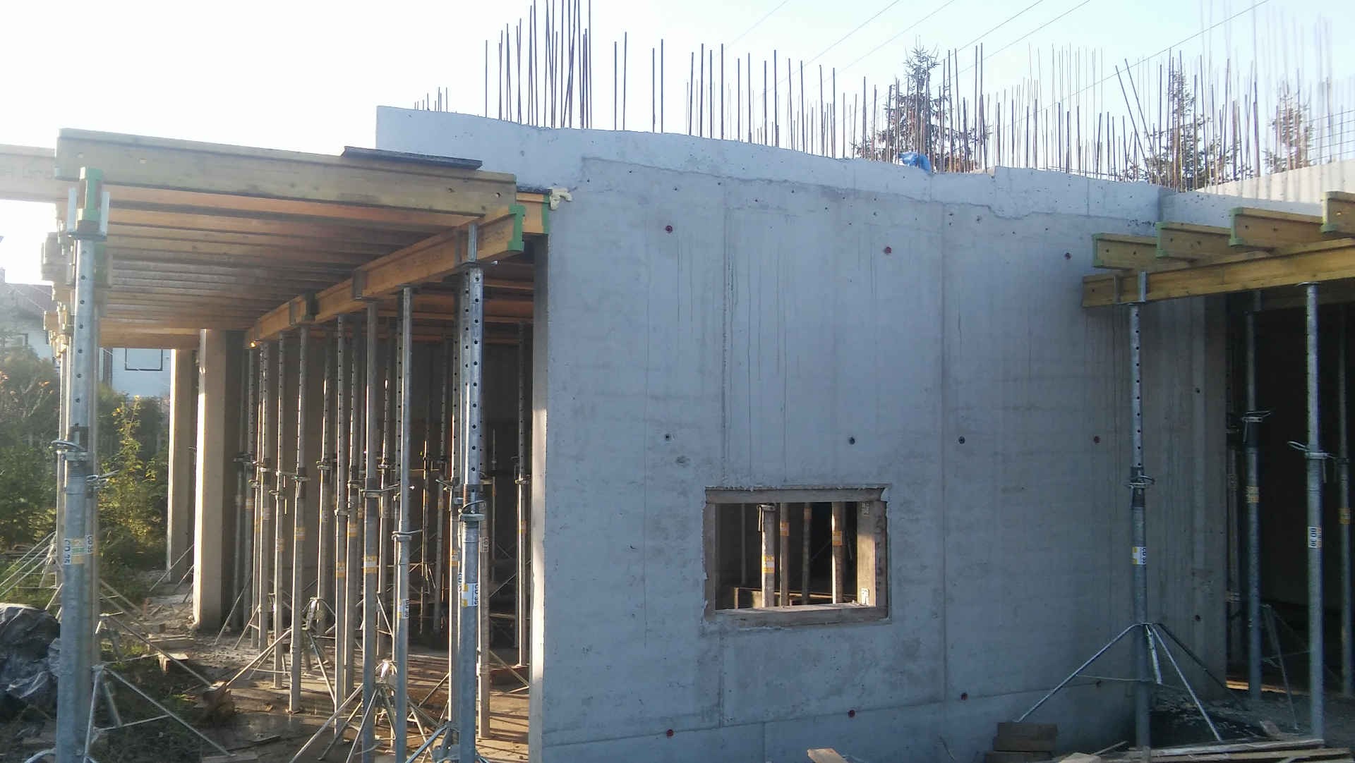 Surowa betonowa konstrukcja domu w trakcie budowy, widoczne szalunki i stalowe podpory, otwór okienny w ścianie, pręty zbrojeniowe wystające z góry.