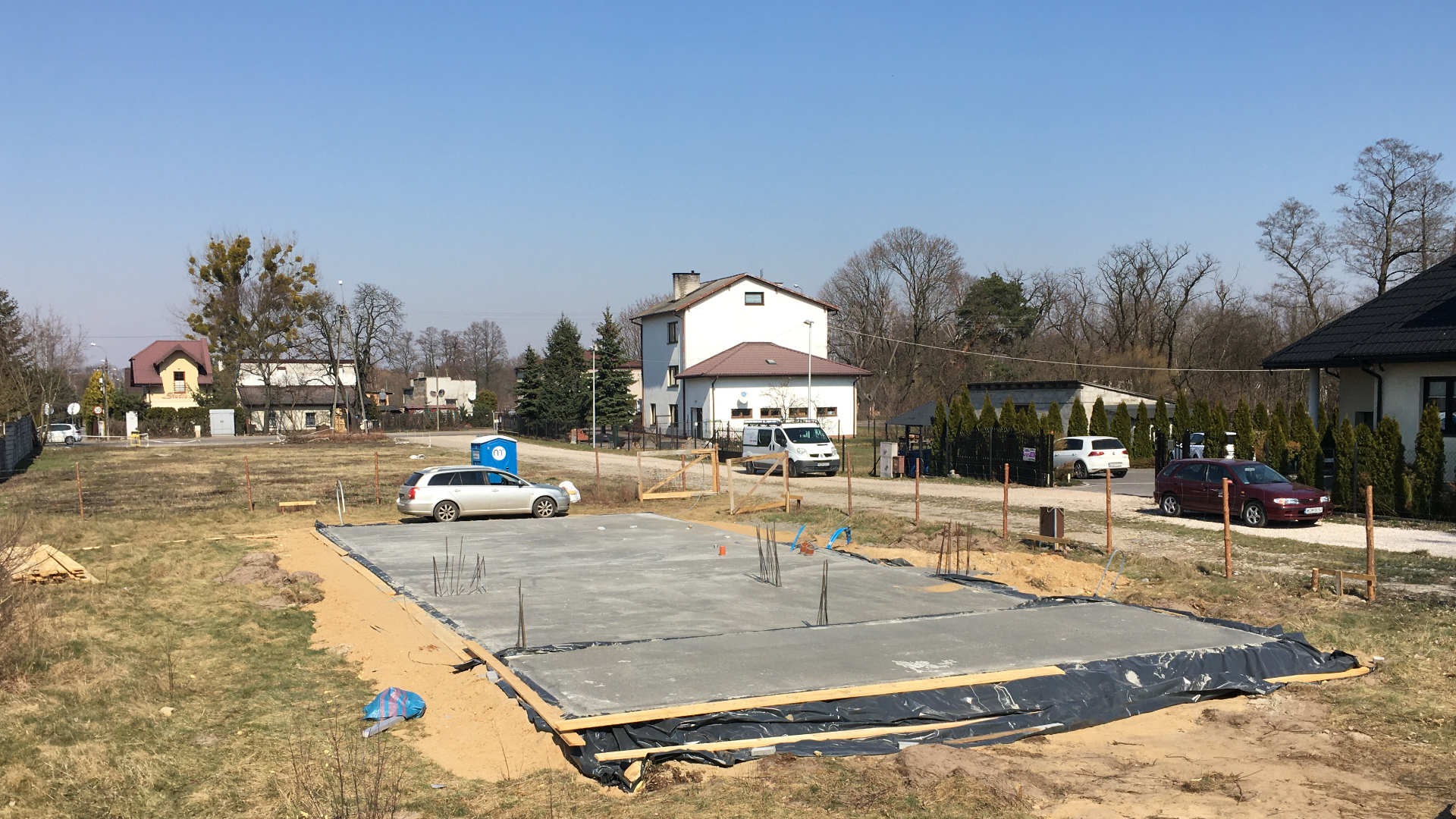 Wylany betonowy fundament pod budowę domu jednorodzinnego na działce, widoczne zbrojenia i szalunki, w tle zabudowa jednorodzinna i błękitne przenośne WC.