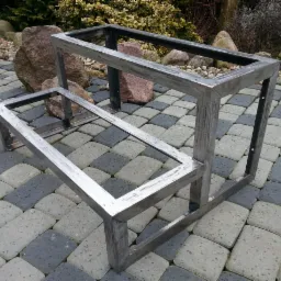 Metalowa konstrukcja altany lub podestu, spawana, z widocznymi śladami obróbki, ustawiona na kostce brukowej w otoczeniu kamieni i roślin.