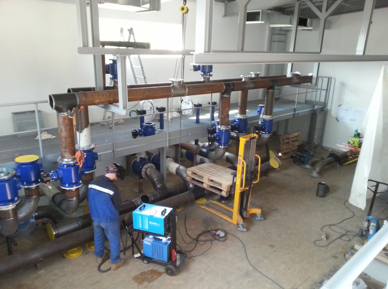 Instalacja przemysłowa z rur i zaworów, spawacz w niebieskim kombinezonie i masce spawalniczej przy pracy, widoczny wózek widłowy z paletą, elementy konstrukcji stalowej pod sufitem.