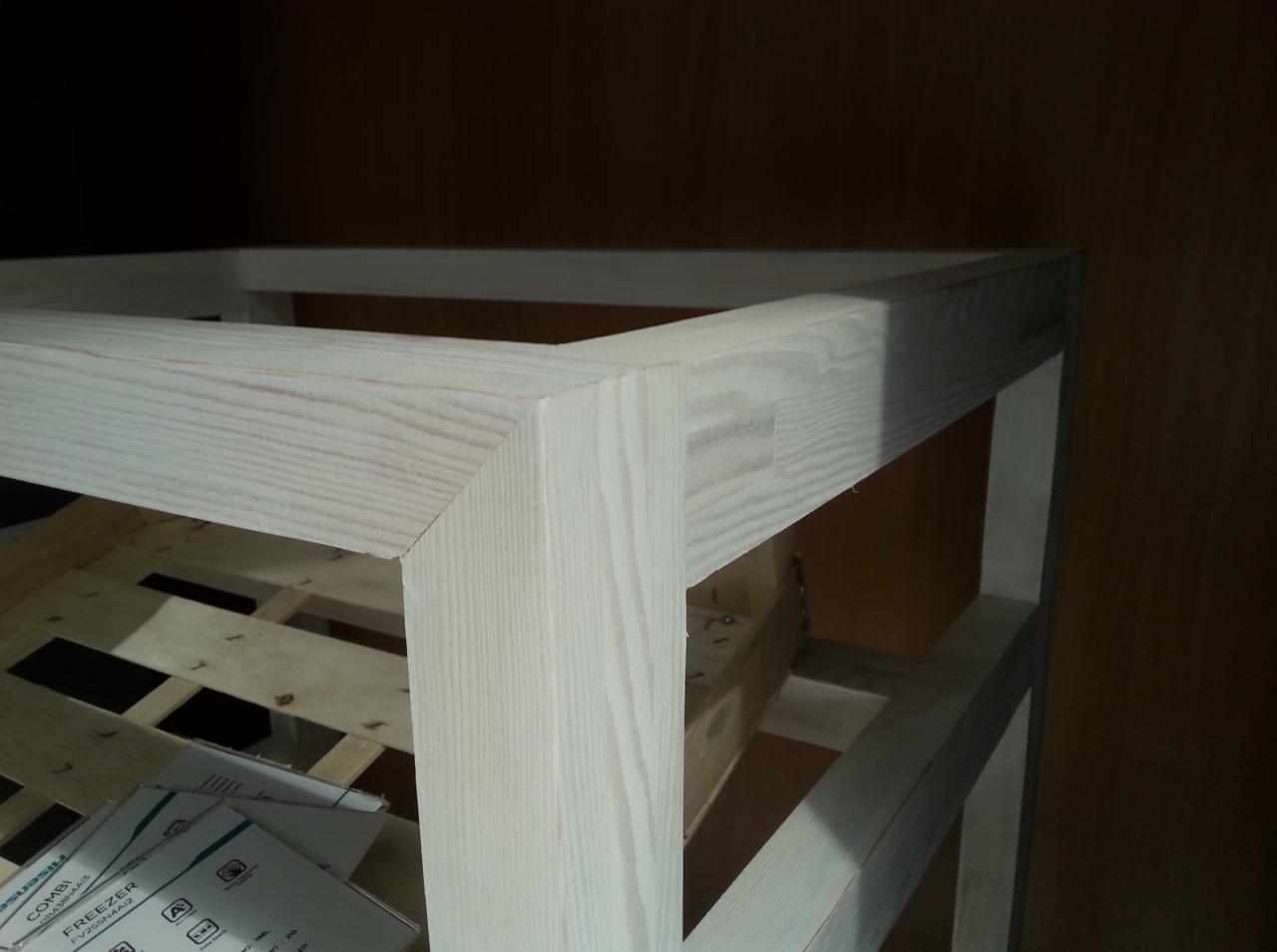 Fragment drewnianej konstrukcji o jasnym odcieniu, z widocznymi połączeniami elementów i widoczną strukturą drewna, oświetlona z góry, z elementami opakowania w tle.