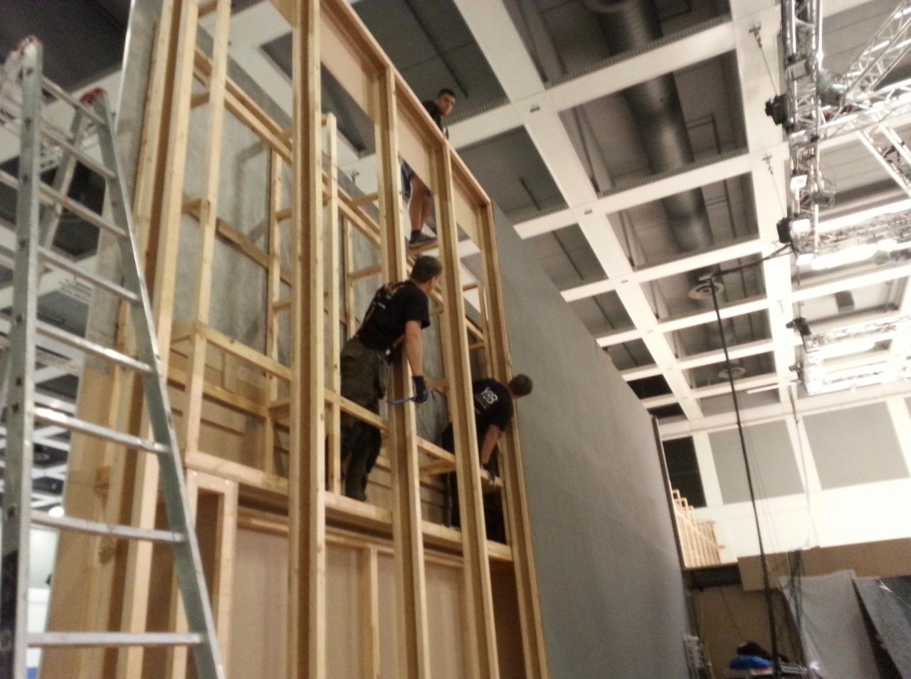 Montaż drewnianej konstrukcji ścian działowych w hali, trzech pracowników w czarnych koszulkach przy pracy, widoczna drabina aluminiowa i oświetlenie sufitowe.