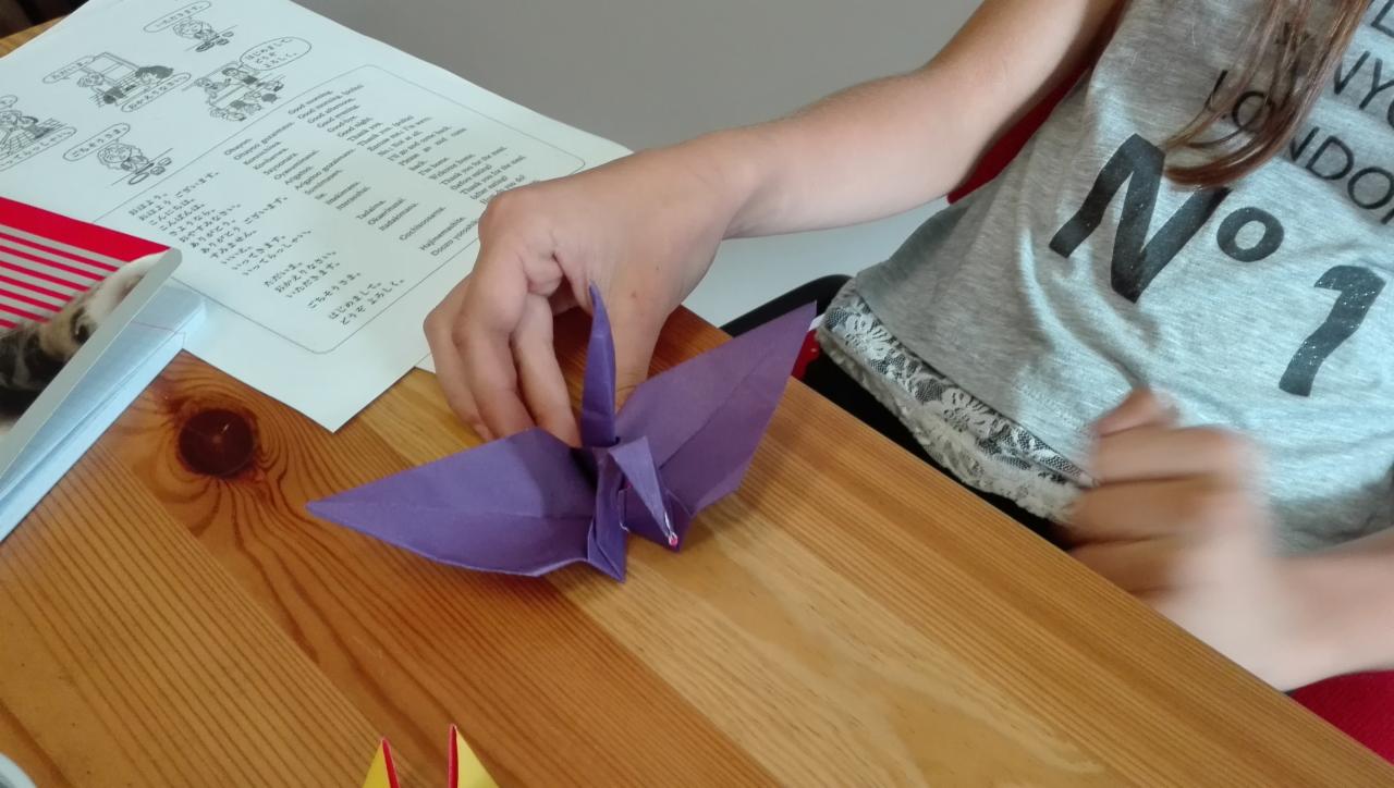 Dziewczynka składa origami żurawia z fioletowego papieru na drewnianym stole, obok leży kartka z tekstem w języku japońskim i fragment origami, kot spogląda zza kartki.