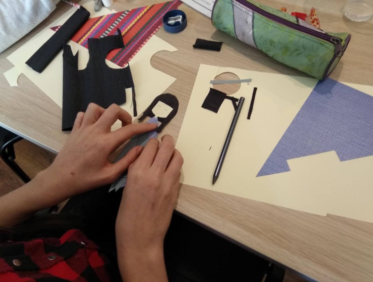 Ręce składają papierową figurkę kobiety z wyciętych elementów na biurku, obok leży piórnik i inne materiały plastyczne.