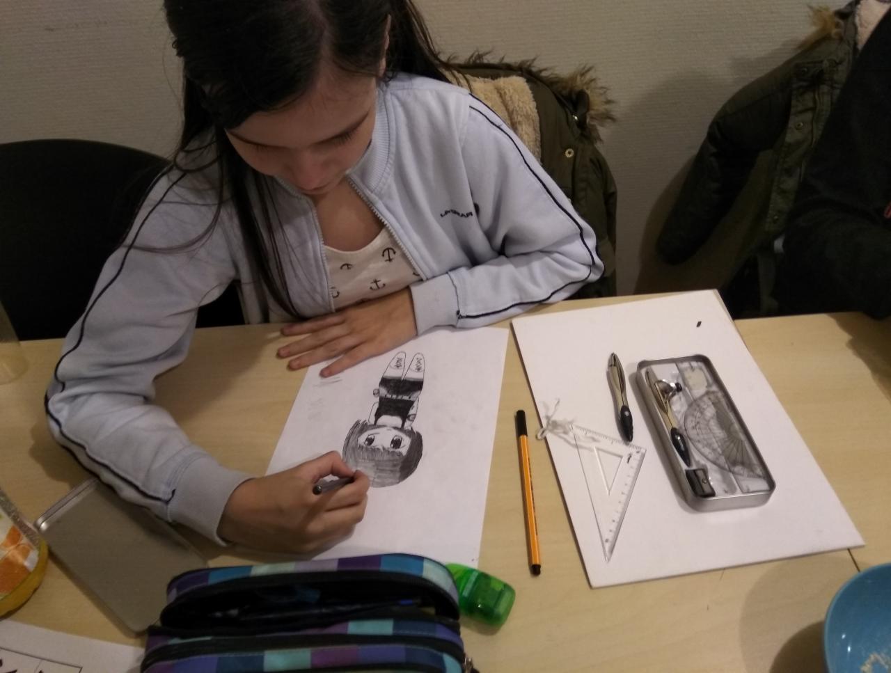 Dziewczynka rysuje mangę na kartce, używając czarnego pisaka. Na biurku leżą przybory geometryczne, piórnik i telefon.