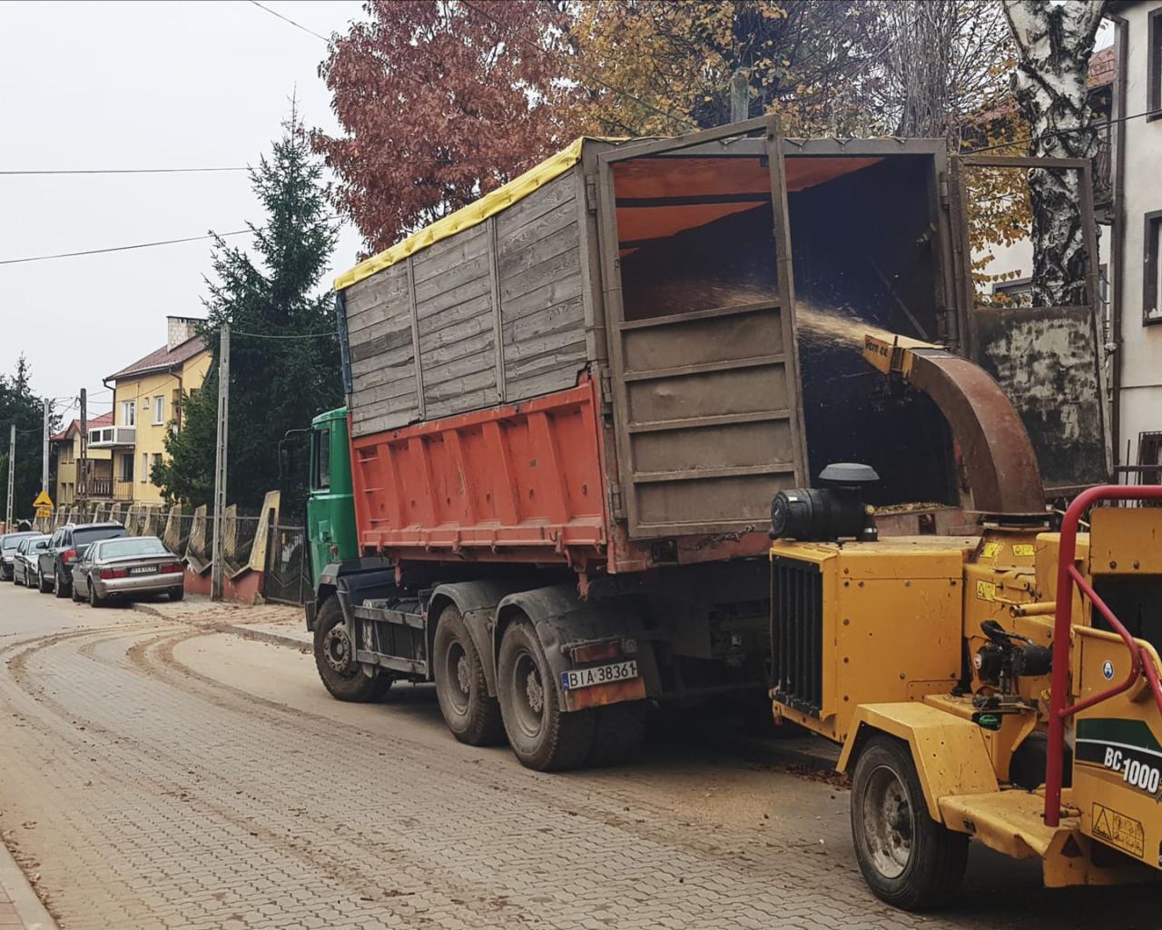 Żółty rębak do gałęzi podaje rozdrobnione drewno do skrzyni ładunkowej ciężarówki na tle ulicy z zaparkowanymi samochodami i budynkami.