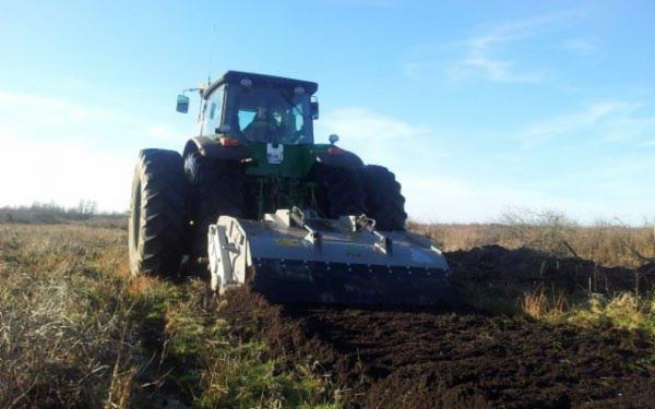 Ciężki traktor z glebogryzarką na polu, świeżo spulchniona ziemia, niebieskie niebo w tle.