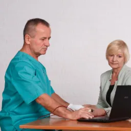 Lekarz w zielonym uniformie podłącza elektrody do nadgarstka pacjentki podczas diagnostyki przy użyciu laptopa na drewnianym stole, na tle białej ściany z cegieł.