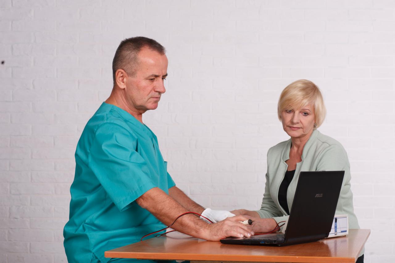 Lekarz w zielonym uniformie podłącza elektrody do nadgarstka pacjentki podczas diagnostyki przy użyciu laptopa na drewnianym stole, na tle białej ściany z cegieł.