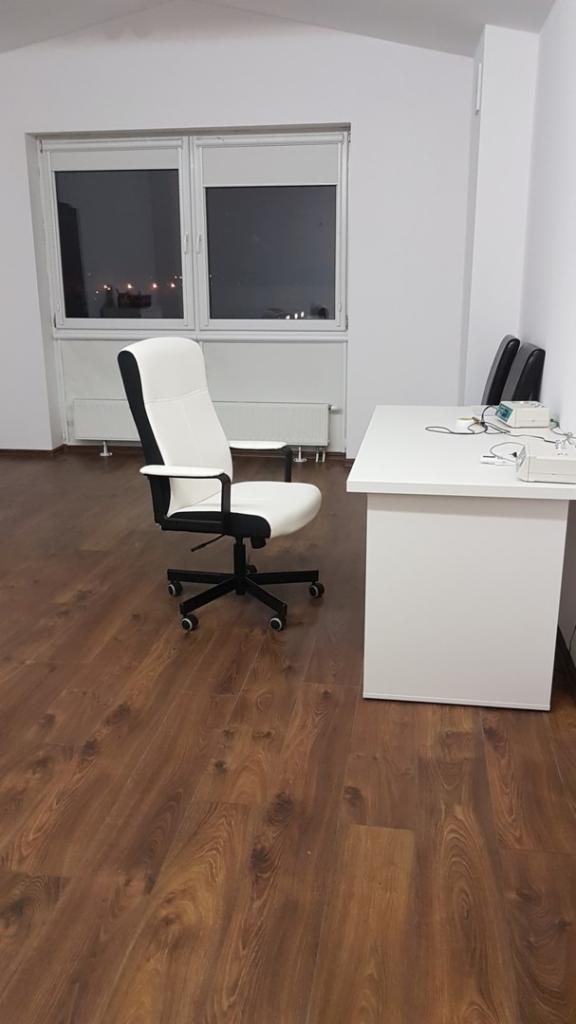 Gabinet z białym biurkiem, krzesłem biurowym w kolorze białym i czarnym oraz urządzeniami medycznymi na blacie, widok z okna na nocne miasto.