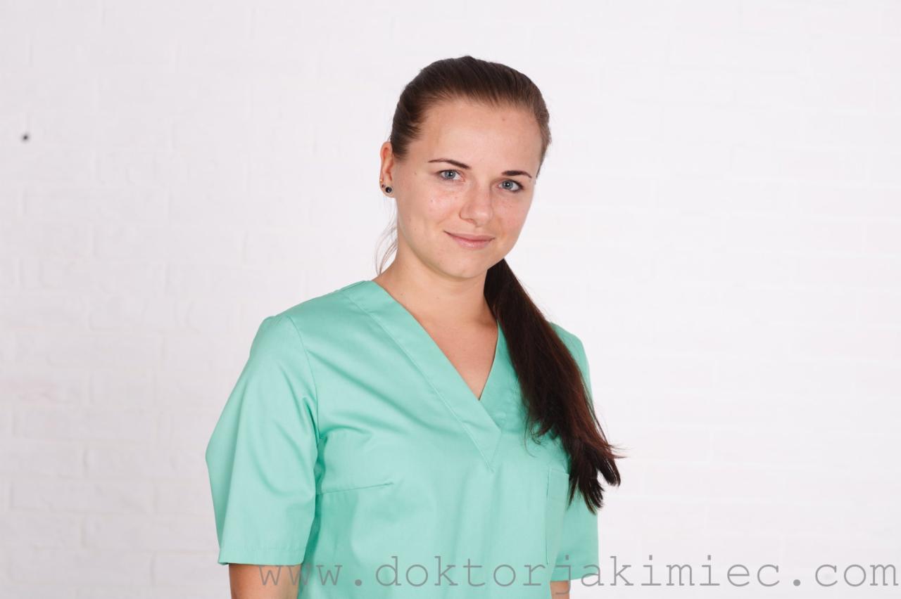 Portret uśmiechniętej kobiety w turkusowym uniformie medycznym na tle białej ściany z cegieł.