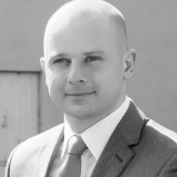 Portret biznesowy mężczyzny w garniturze, z delikatnym uśmiechem, na tle szarej ściany i drzwi, w czerni i bieli.