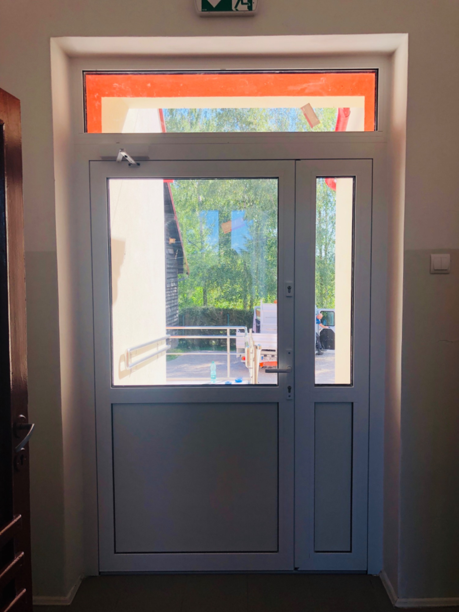 Białe, przeszklone drzwi dwuskrzydłowe z górnym świetlikiem wewnątrz budynku, widok na zewnątrz, gdzie stoi podnośnik koszowy i widać zieleń.