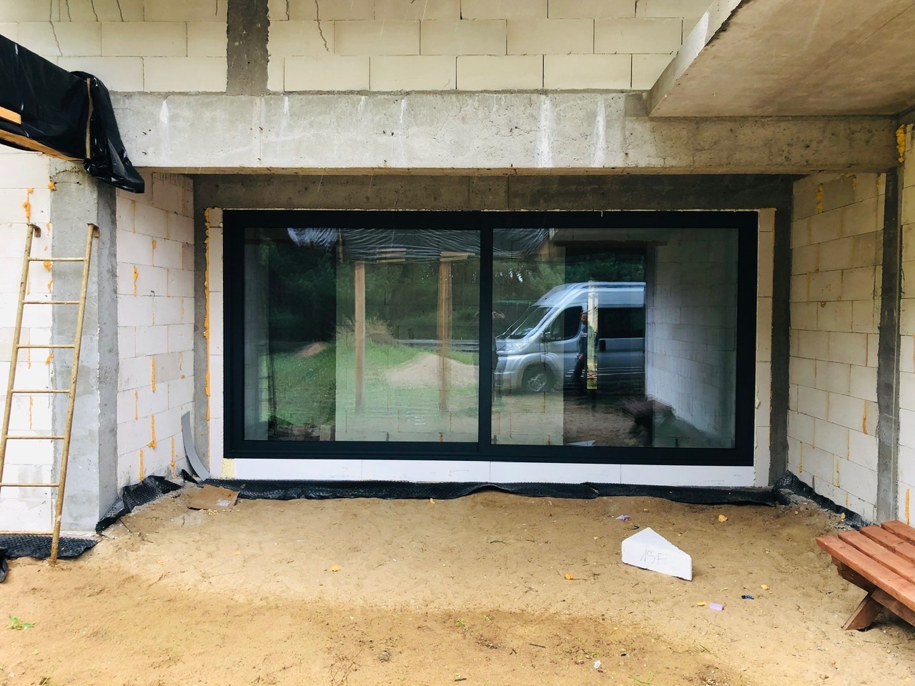 Duże, czarne okno przesuwne zamontowane w surowym stanie budynku z białego bloczka, widoczny van na zewnątrz odbijający się w szybie, piasek na podłożu i drabina oparta o ścianę.