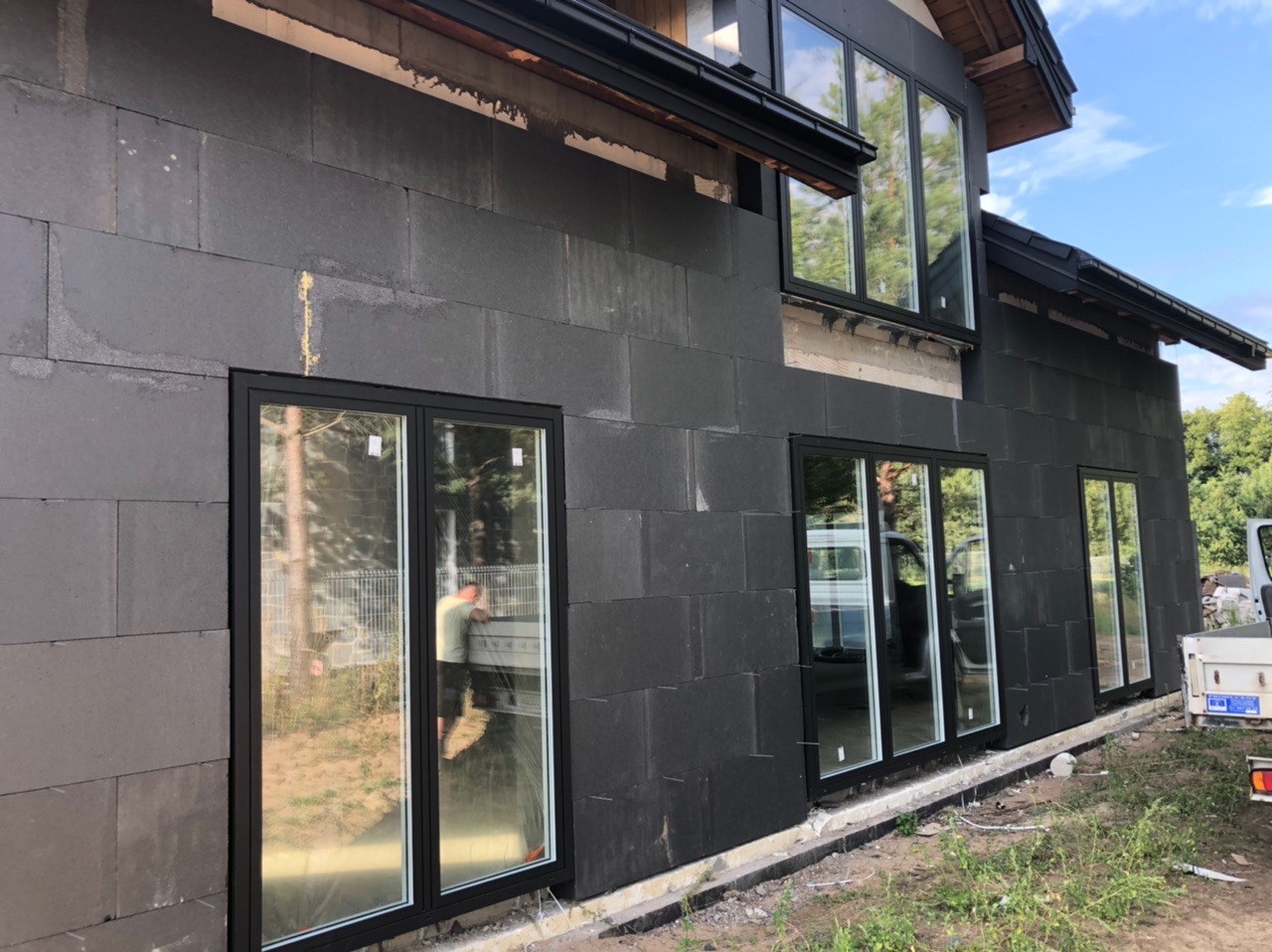 Częściowy widok elewacji domu z szarego betonu komórkowego z zamontowanymi oknami z czarnymi ramami; w szybie jednego okna odbija się sylwetka mężczyzny pracującego przy samochodzie dostawczym.