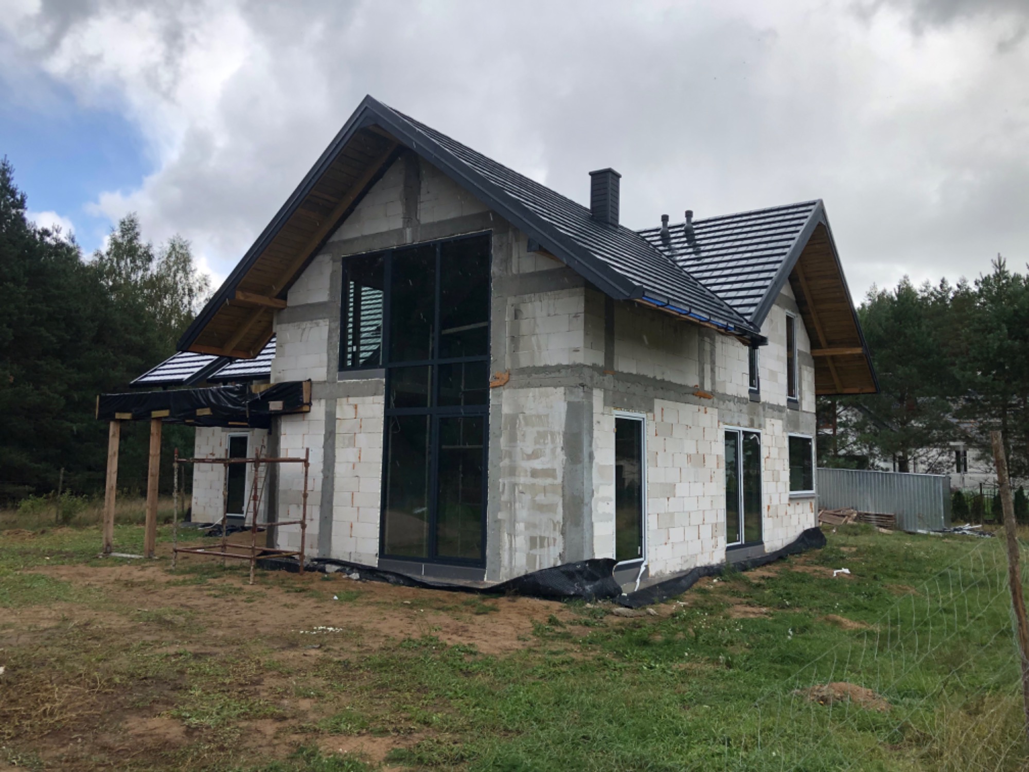 Surowy stan domu w trakcie budowy z widocznymi oknami z czarnymi ramami i niezabezpieczonymi otworami drzwiowymi, na tle pochmurnego nieba i drzew