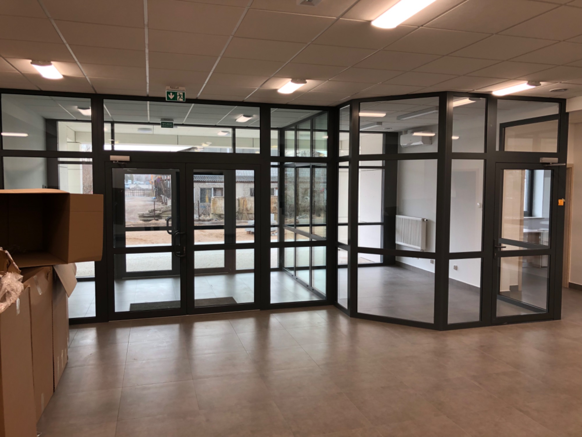 Szklana ścianka działowa z drzwiami w nowym biurze, widoczne kartony po lewej stronie, płytki podłogowe w odcieniach beżu.