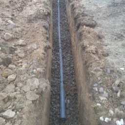 Wykop w ziemi z ułożoną rurą drenażową obsypaną drobnymi kamieniami, widoczne ślady świeżo wykopanej ziemi. Rura szara, plastikowa, ułożona wzdłuż wykopu.