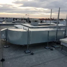 Metalowy kanał wentylacyjny o zaokrąglonym kształcie, wsparty na metalowych nogach z czarnymi podstawami, umieszczony na płaskim dachu budynku wielorodzinnego z widokiem na inne budynki...