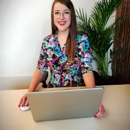 Uśmiechnięta kobieta w okularach i wzorzystej sukience siedzi za biurkiem z laptopem i myszką, w tle roślina doniczkowa i drewniana ściana.