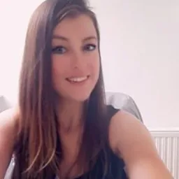 Portret uśmiechniętej brunetki z długimi włosami, w czarnej bluzce, na jasnym tle. Kadr od ramion w górę, delikatny uśmiech, naturalny makijaż.