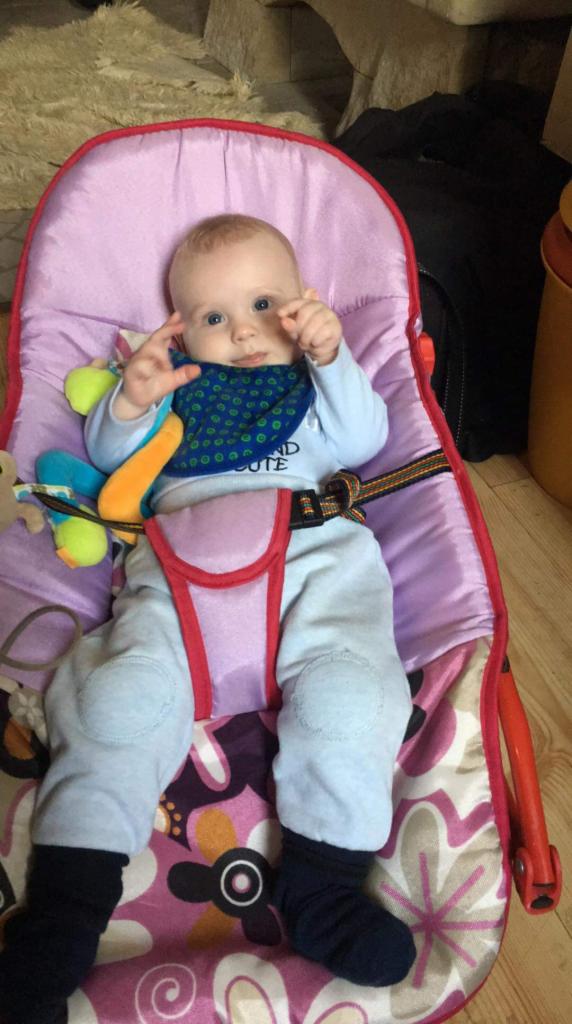 Niemowlę z niebieskimi oczami siedzi w różowo-czerwonym leżaczku z pasami, ubrane w niebieską bluzkę z napisem i szare spodnie, na podłodze wzorzysty dywan.