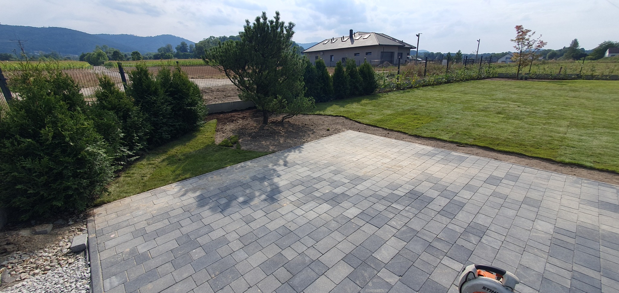Widok z góry na świeżo ułożony, szary chodnik z kostki brukowej, przylegający do zadbanego trawnika z nasadzeniami tuj i drzew, z widokiem na dom i górzysty krajobraz w tle.