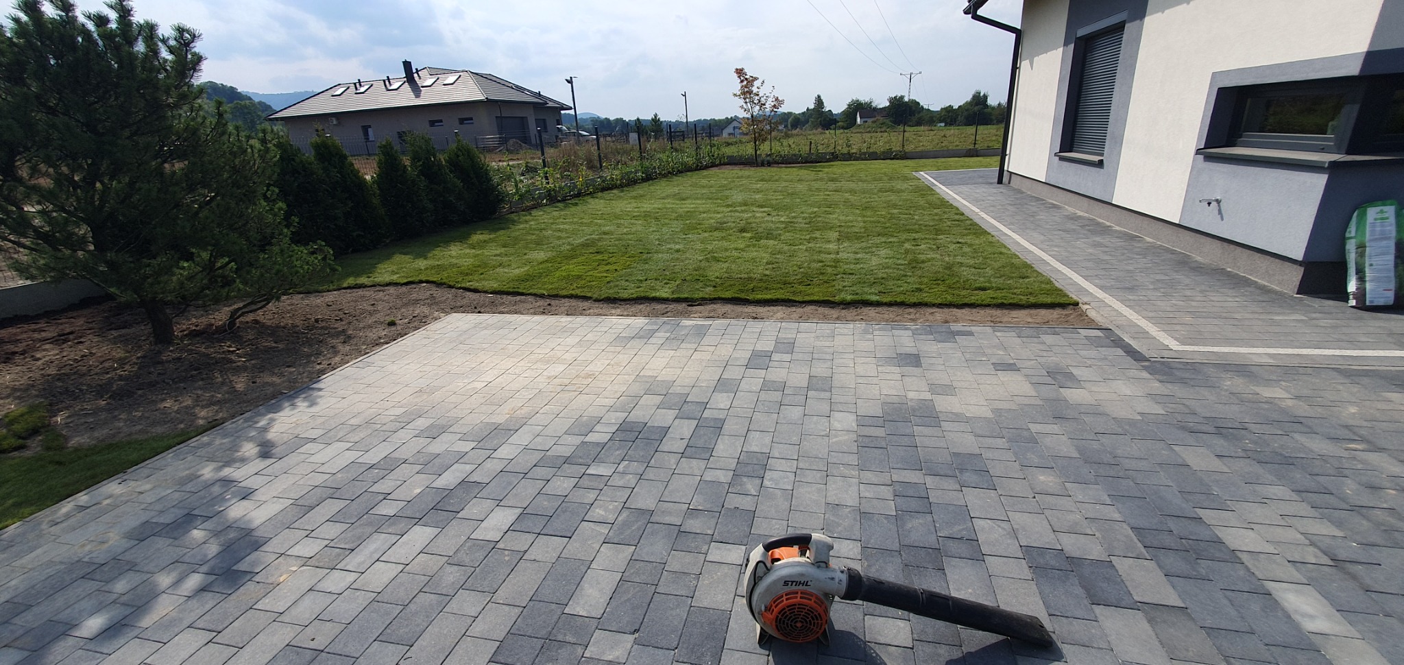 Świeżo założony trawnik przylegający do domu z szarą kostką brukową, na której leży dmuchawa do liści Stihl. W tle widoczny dom jednorodzinny i zieleń.