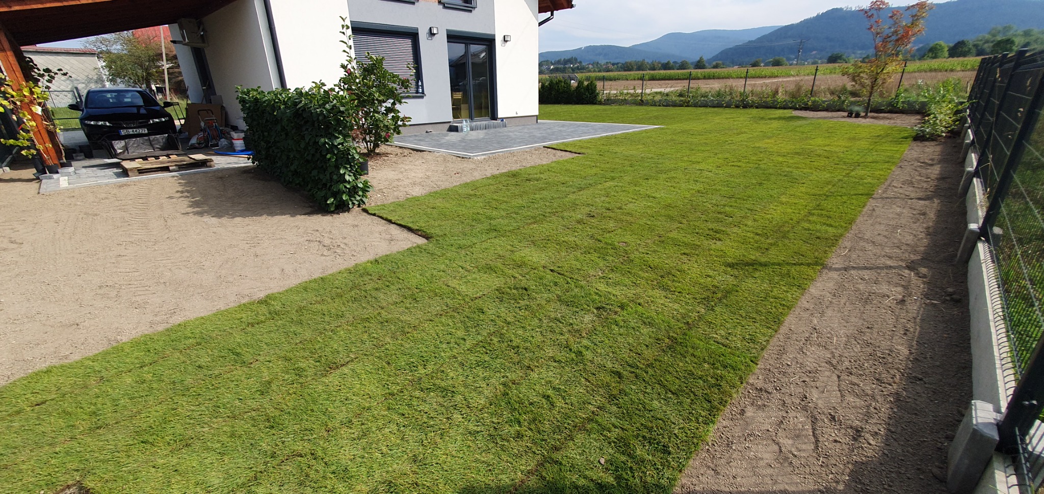Widok na świeżo rozłożony trawnik z rolki, przylegający do domu z szarą elewacją i kostką brukową, z widokiem na góry w tle.