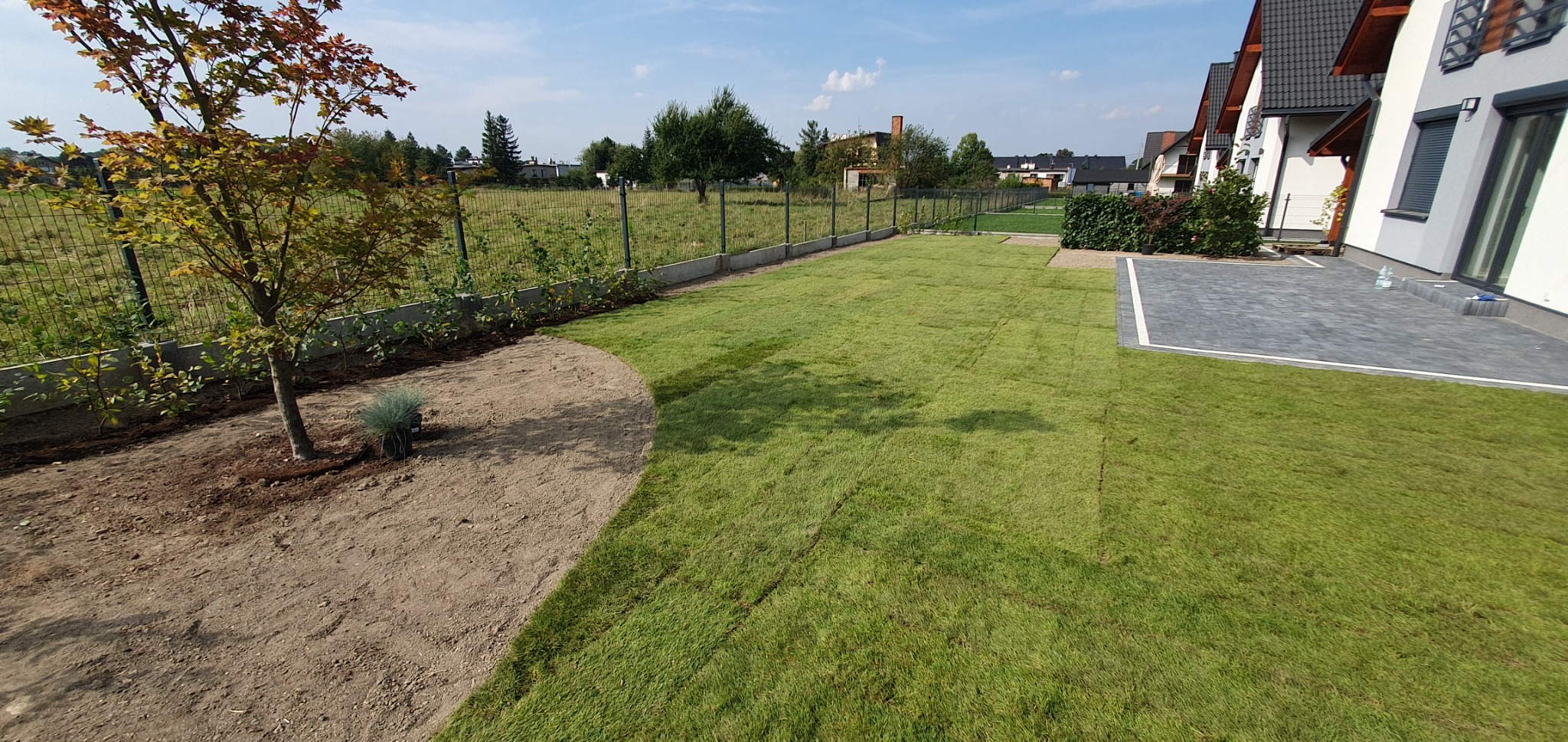 Widok z góry na świeżo rozłożony trawnik z rolki, z widocznymi liniami łączeń, graniczący z przygotowanym podłożem pod nasadzenia i kostką brukową przy domu.