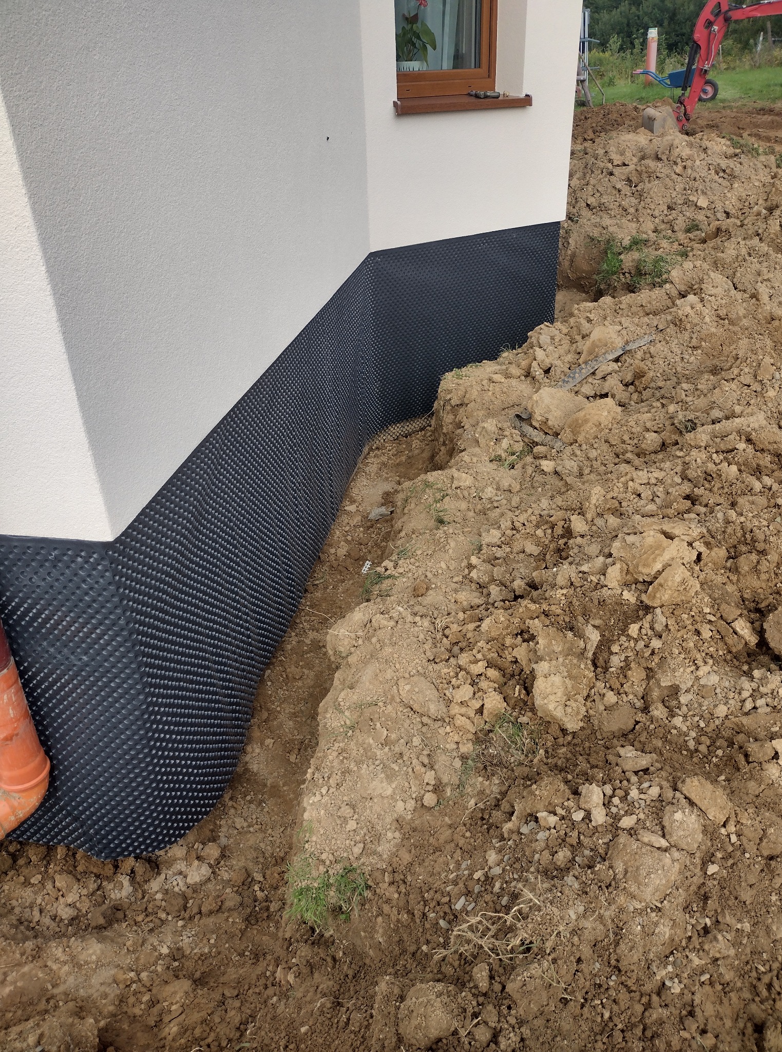 Ochrona fundamentów budynku folią kubełkową i wykop ziemny. W tle widoczny minikoparka, okno budynku. Prace ziemne w toku.