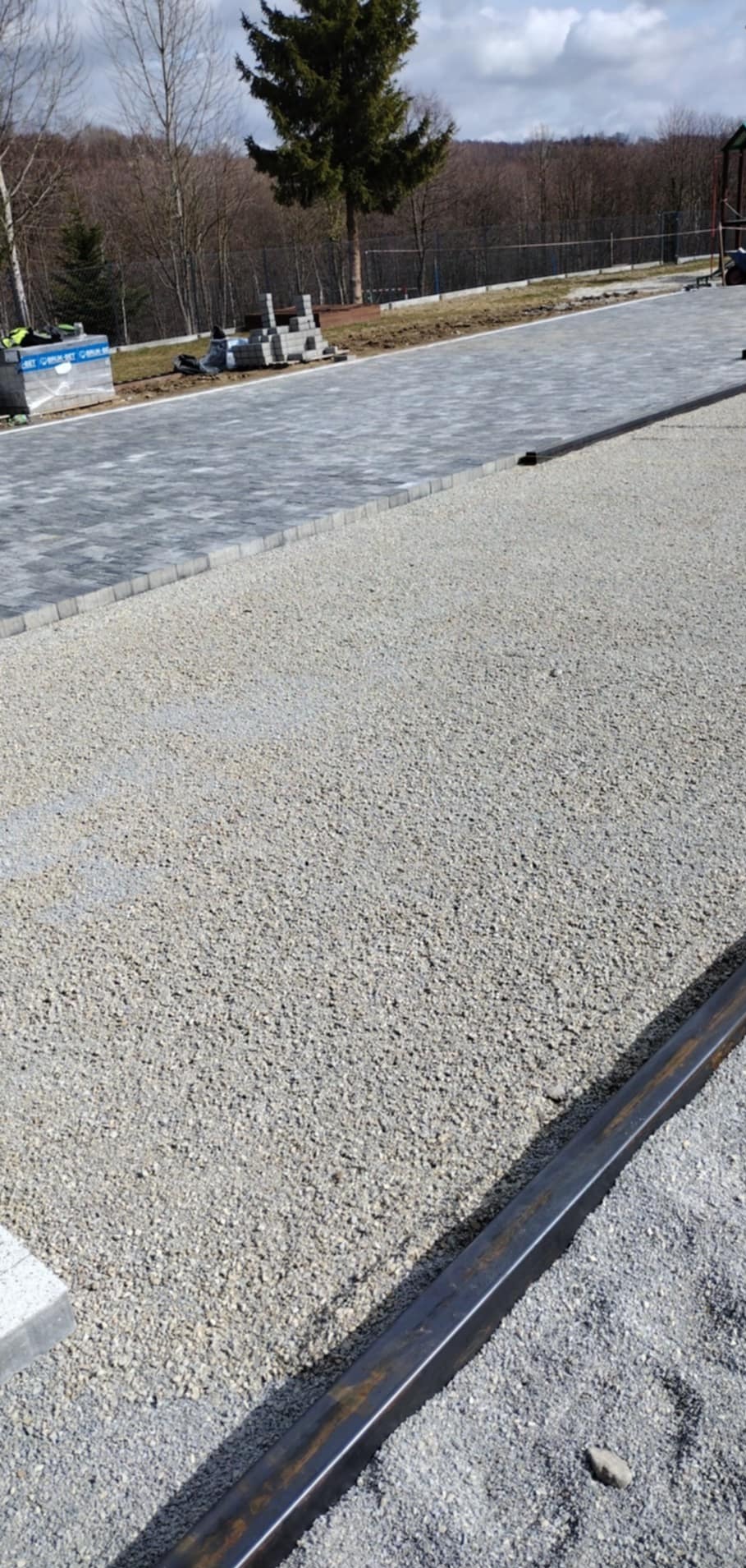 Etap przygotowania podłoża pod kostkę brukową: wysypany żwir, widoczne szyny prowadzące. W tle ułożona część kostki i zapas materiału. Dzień.