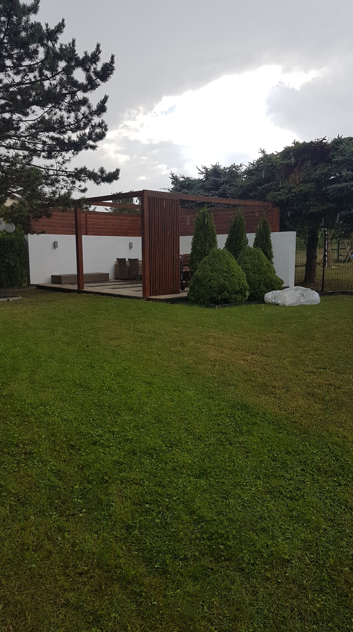 Nowoczesna pergola z drewnianymi elementami i białymi ścianami w zielonym ogrodzie. Widoczne krzewy i drzewa iglaste, oświetlenie zewnętrzne.