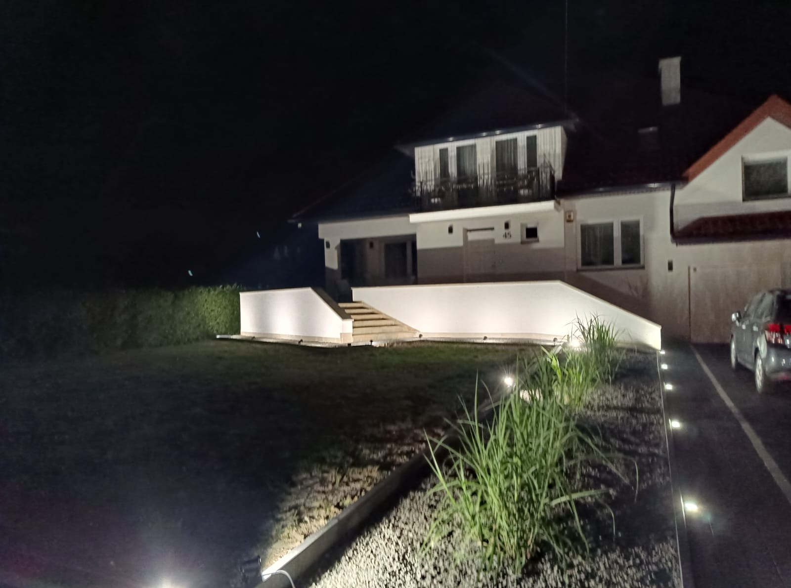 Nowoczesny dom z podświetloną elewacją i ogrodem w nocy. Widoczne schody, trawnik, rośliny ozdobne i fragment samochodu. Oświetlenie podkreśla architekturę budynku.