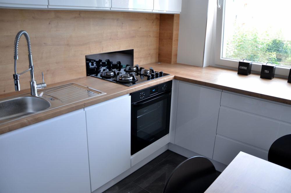 Nowoczesna kuchnia z białymi, lakierowanymi szafkami, drewnianym blatem i zabudowaną płytą grzewczą z piekarnikiem, uzupełniona o czarne pojemniki na kawę i herbatę przy oknie.
