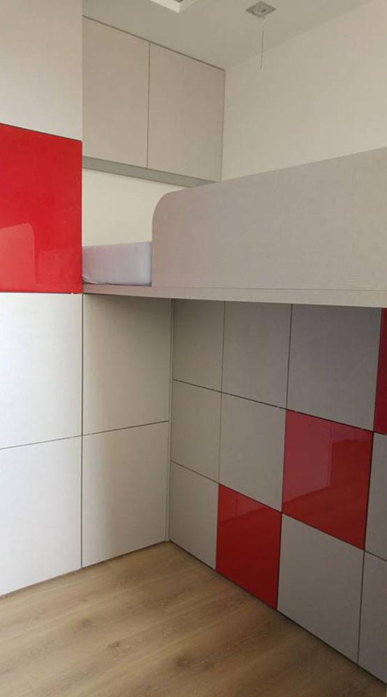 Wnętrze z zabudową szaf i półek w kolorach białym, szarym i czerwonym, tworzących geometryczny wzór. Nad szafami znajduje się antresola z materacem.