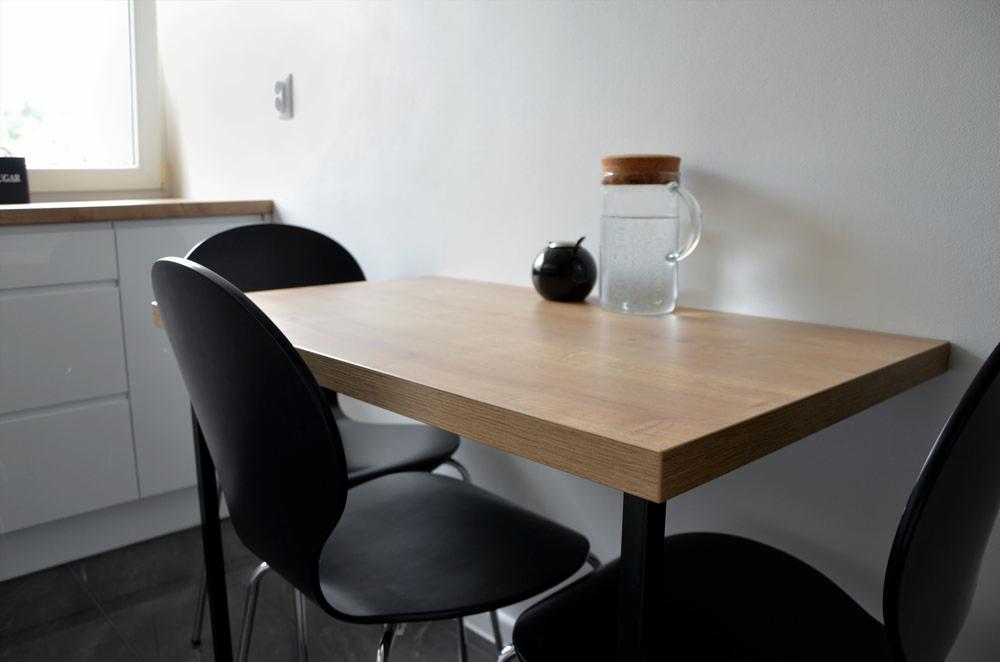 Stół z blatem drewnianym na czarnych nogach, otoczony czarnymi krzesłami, na blacie dzbanek z wodą i czarny pojemnik, w tle białe ściany i zabudowa kuchenna.