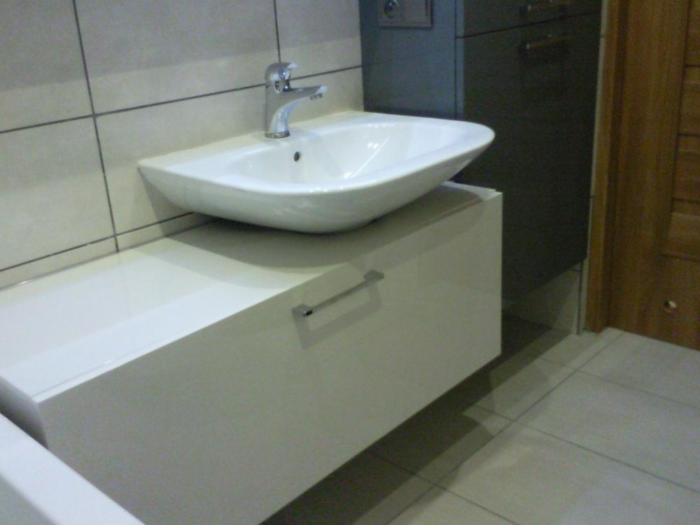 Biała umywalka nablatowa z chromowaną baterią, zamontowana na szafce z białym, błyszczącym frontem. W tle widoczna szara szafka i fragment drewnianych drzwi.