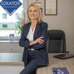 Lokator Agencja Nieruchomości Anna Bujoczek - Profesjonalny portret kobiety w biurze nieruchomości, w ciemnym garniturze, z logo firmy w tle i na biurku. Uśmiechnięta, pewna siebie.