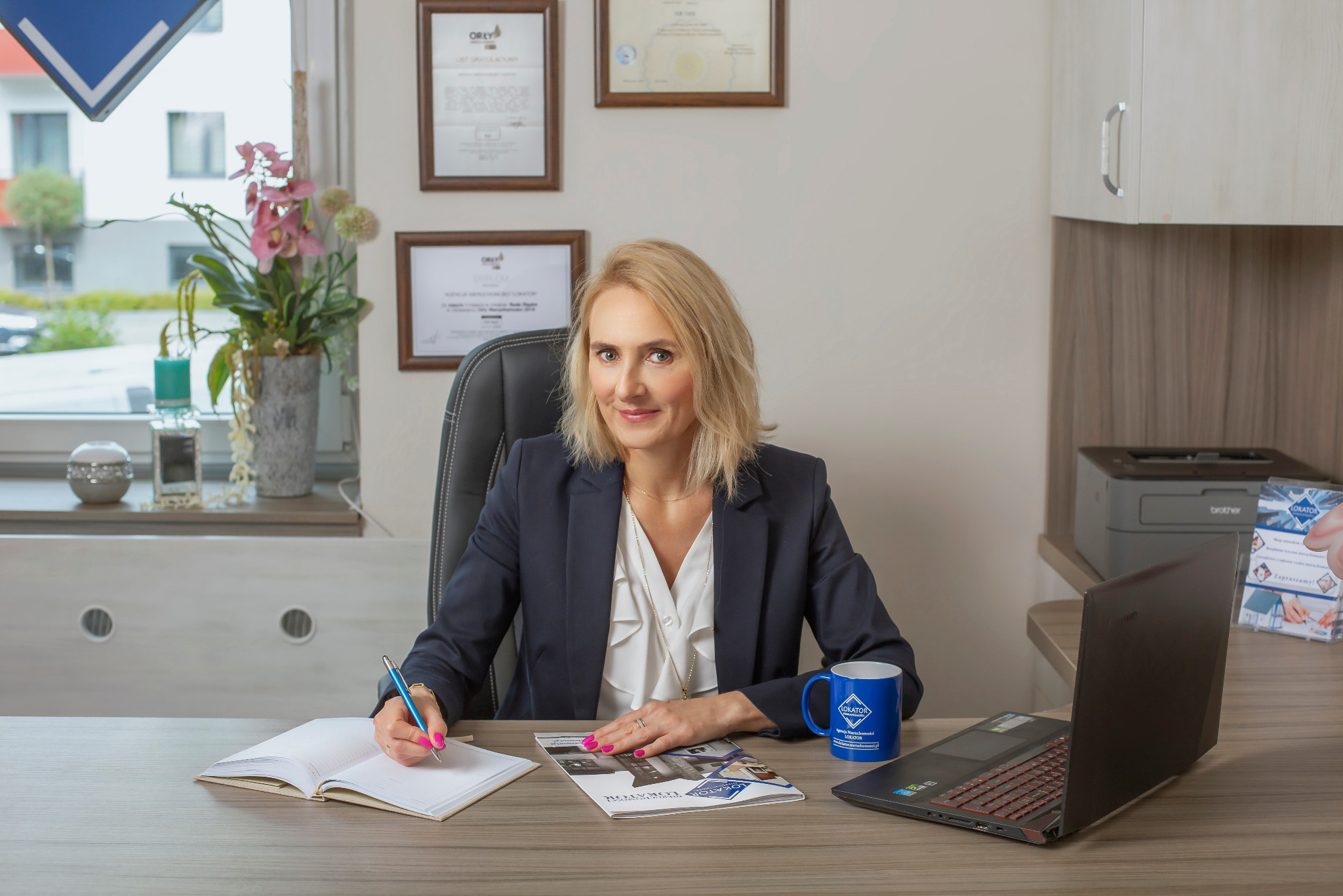 Kobieta w biurze, w ciemnej marynarce, siedzi za biurkiem i pisze w notatniku. Na biurku leży laptop, katalog nieruchomości i niebieski kubek z logo agencji.