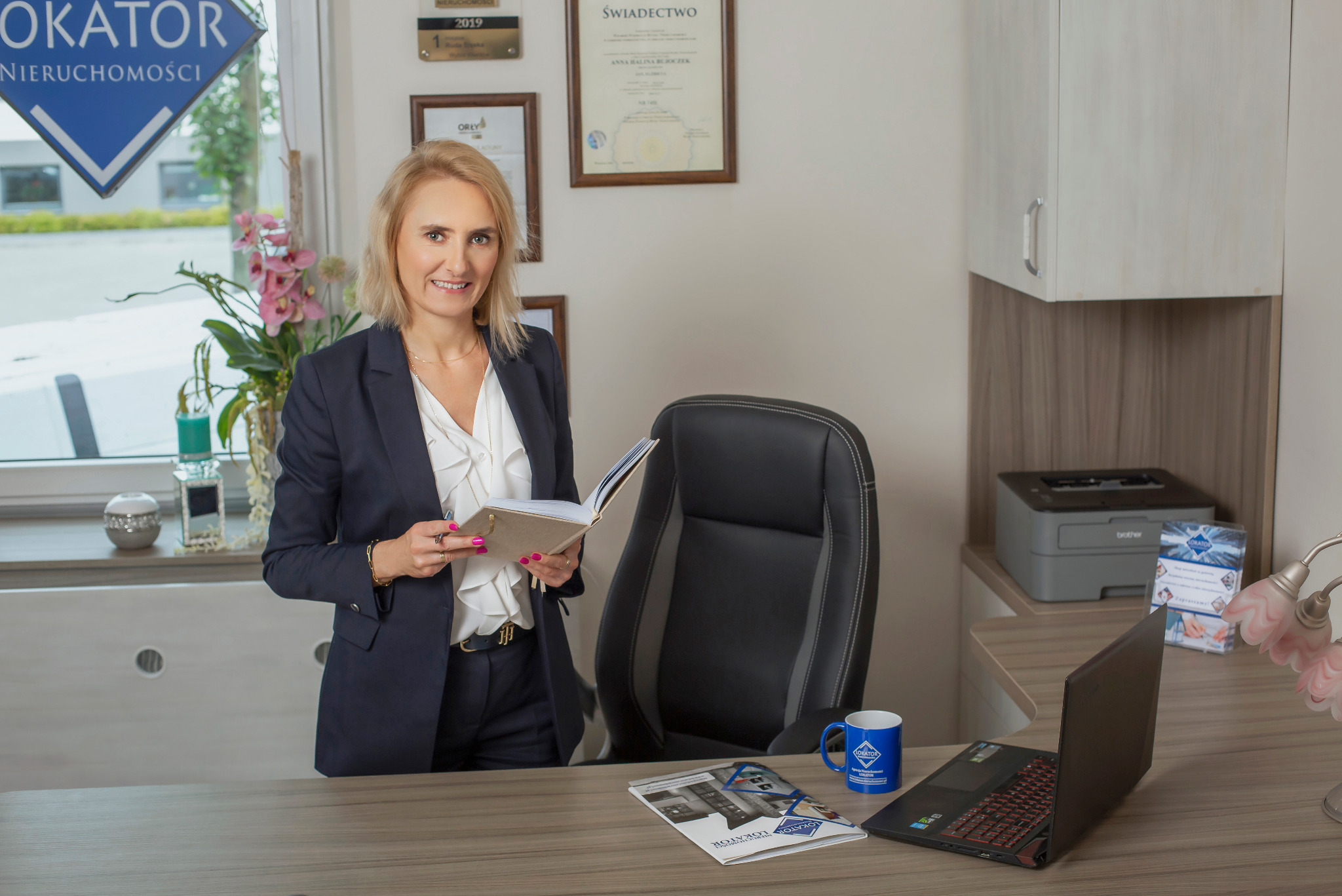 Profesjonalna agentka nieruchomości w biurze, trzymająca notatnik, z logo firmy na kubku i materiałach reklamowych na biurku. Świadectwa i nagrody na ścianie.
