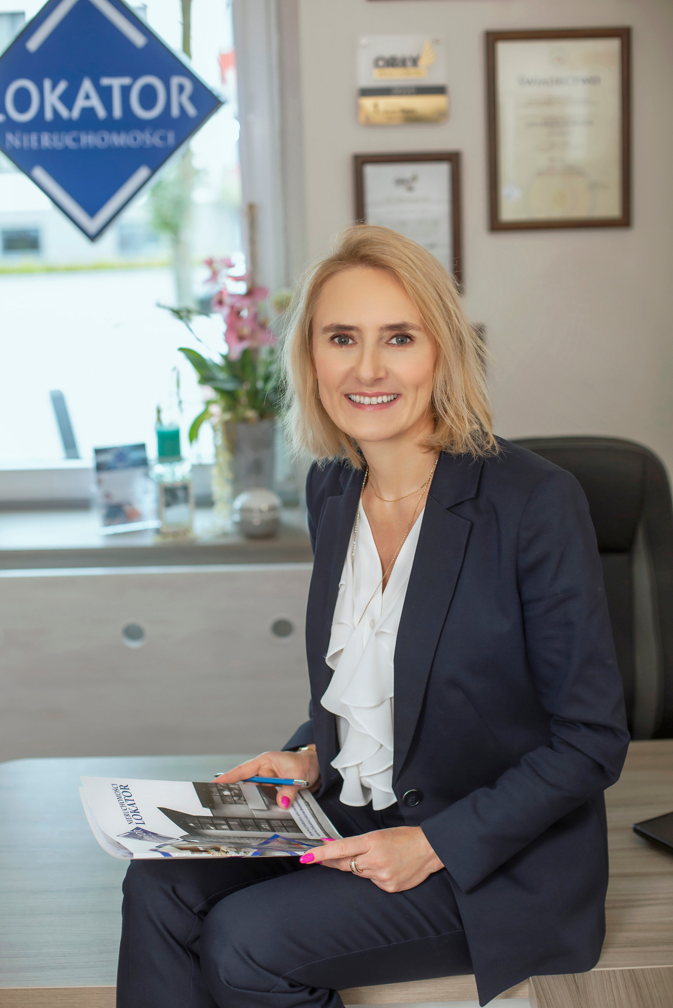 Uśmiechnięta blondynka w biurze nieruchomości 'Lokator', trzymająca folder z ofertami. W tle dyplomy i logo firmy.