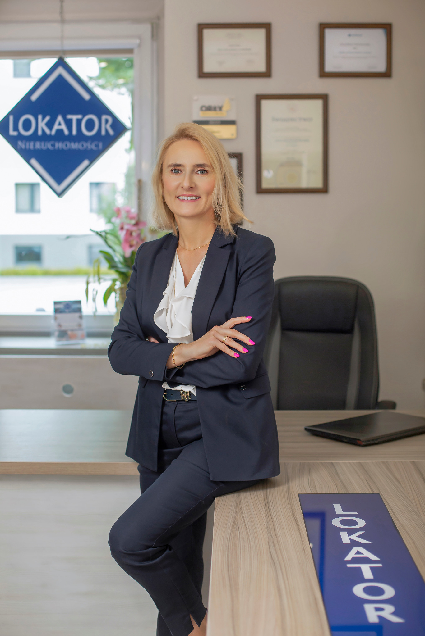 Profesjonalny portret kobiety w biurze nieruchomości, w ciemnym garniturze, z logo firmy w tle i na biurku. Uśmiechnięta, pewna siebie.