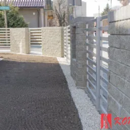 Nowoczesne ogrodzenie posesji z szarego kamienia łupanego i poziomych, metalowych przęseł w kolorze srebrnym, uzupełnione białym grysem.