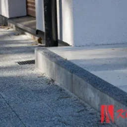 Wykończony chodnik z szarej kostki brukowej z obrzeżem, przylegający do białej ściany budynku z rurą spustową. Widoczna kratka odpływowa i logo firmy w prawym dolnym rogu.