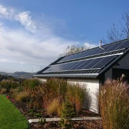 Nowoczesny budynek gospodarczy z panelami słonecznymi na dachu, otoczony ozdobnymi trawami i zadbanym trawnikiem, z widokiem na pagórkowaty krajobraz w tle.